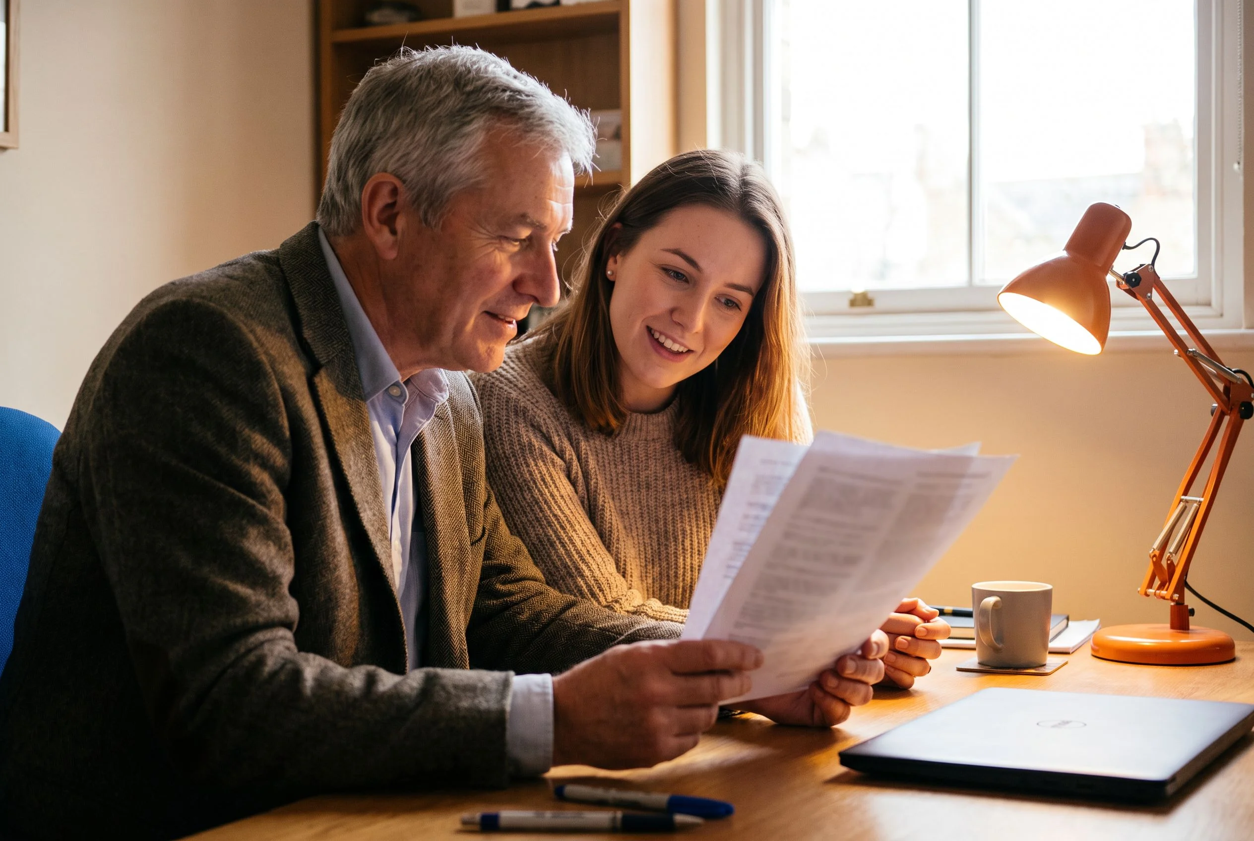 Senior data engineer mentoring a junior colleague reviewing printed documents at a shared workstation, orange accent desk lamp