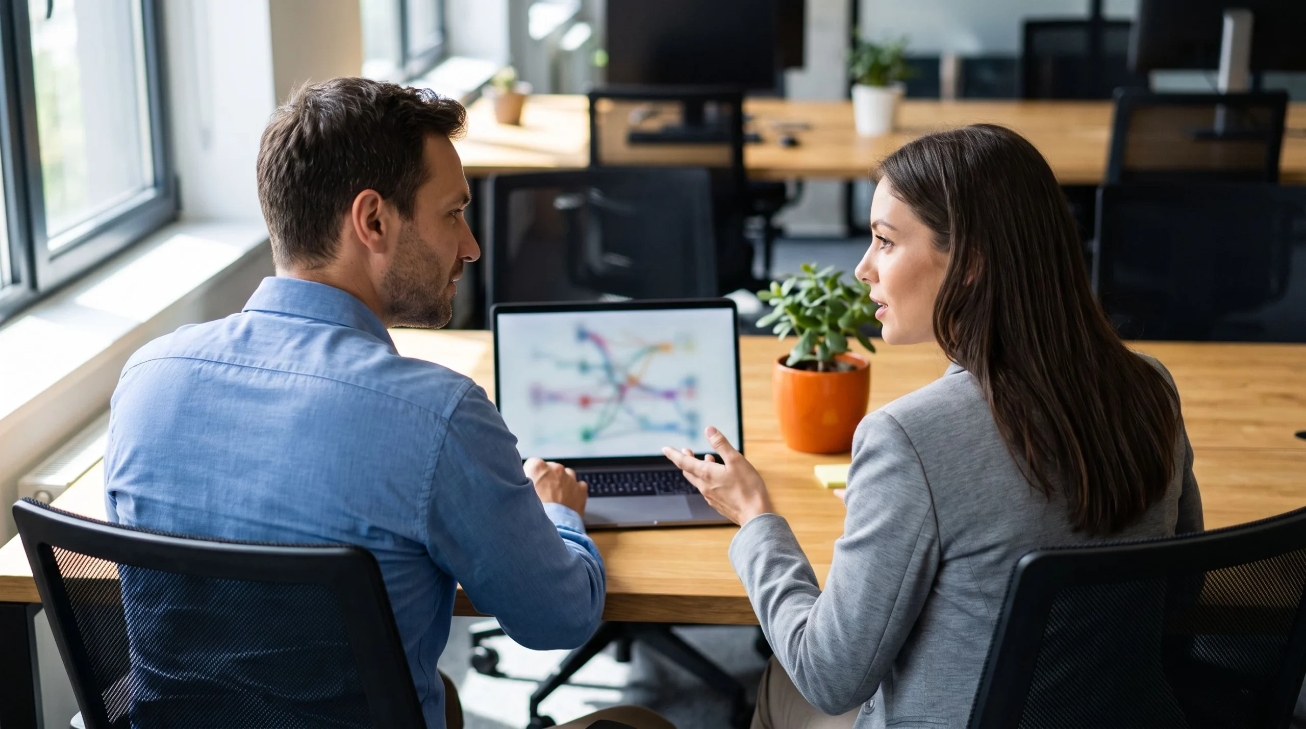New architect being onboarded by a colleague, both looking at a laptop showing a systems diagram, over the shoulder angle, warm orange accent on a desk plant