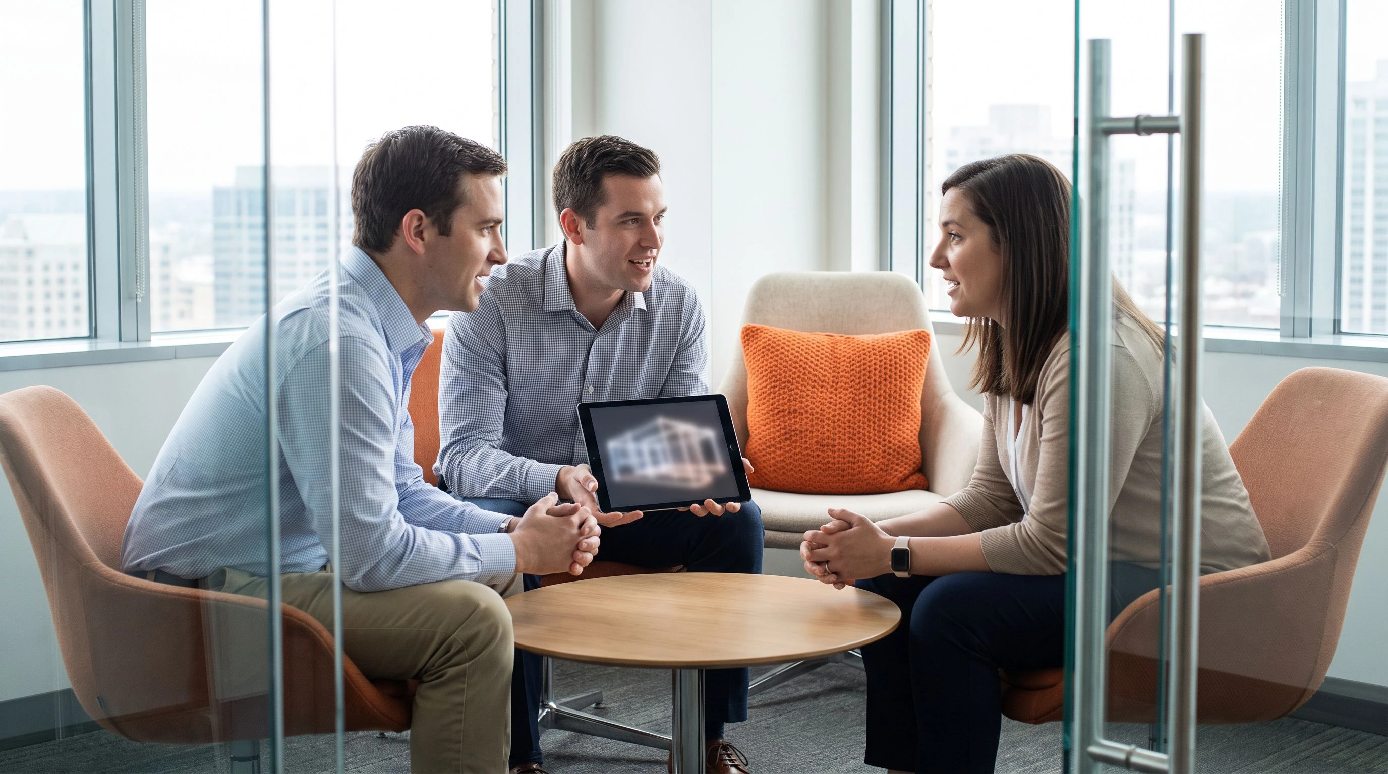 Three professionals in a small architecture review meeting discussing a diagram on a tablet, warm orange accent pillow visible