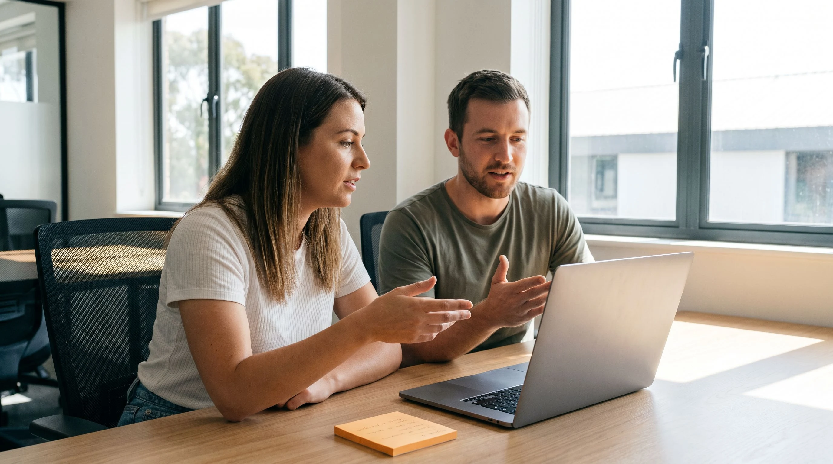 Hiring manager and technical lead reviewing a candidate skills matrix on a laptop screen together, warm orange sticky note visible on the desk