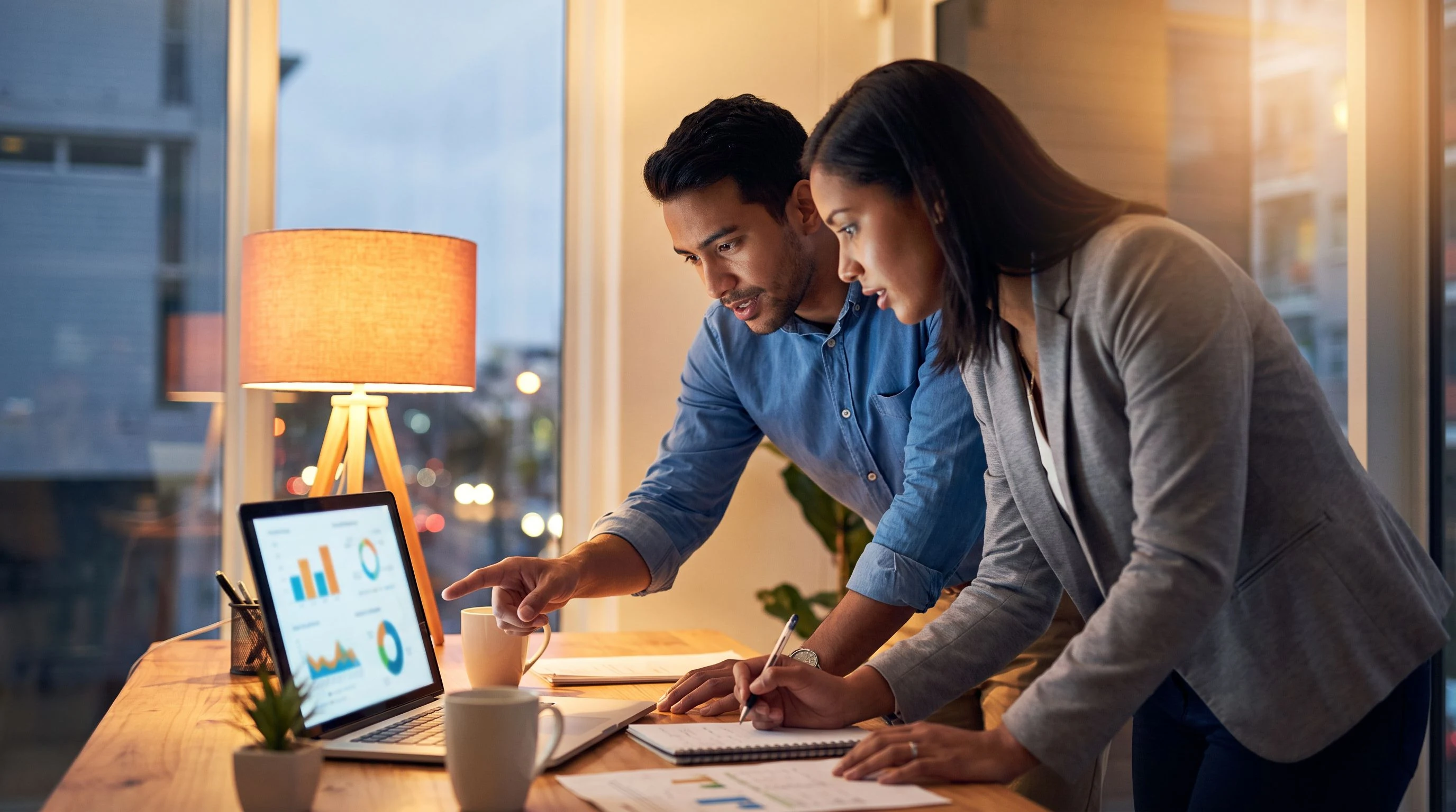 Finance and hiring manager reviewing compensation bands on a laptop at a desk with a warm orange desk lamp