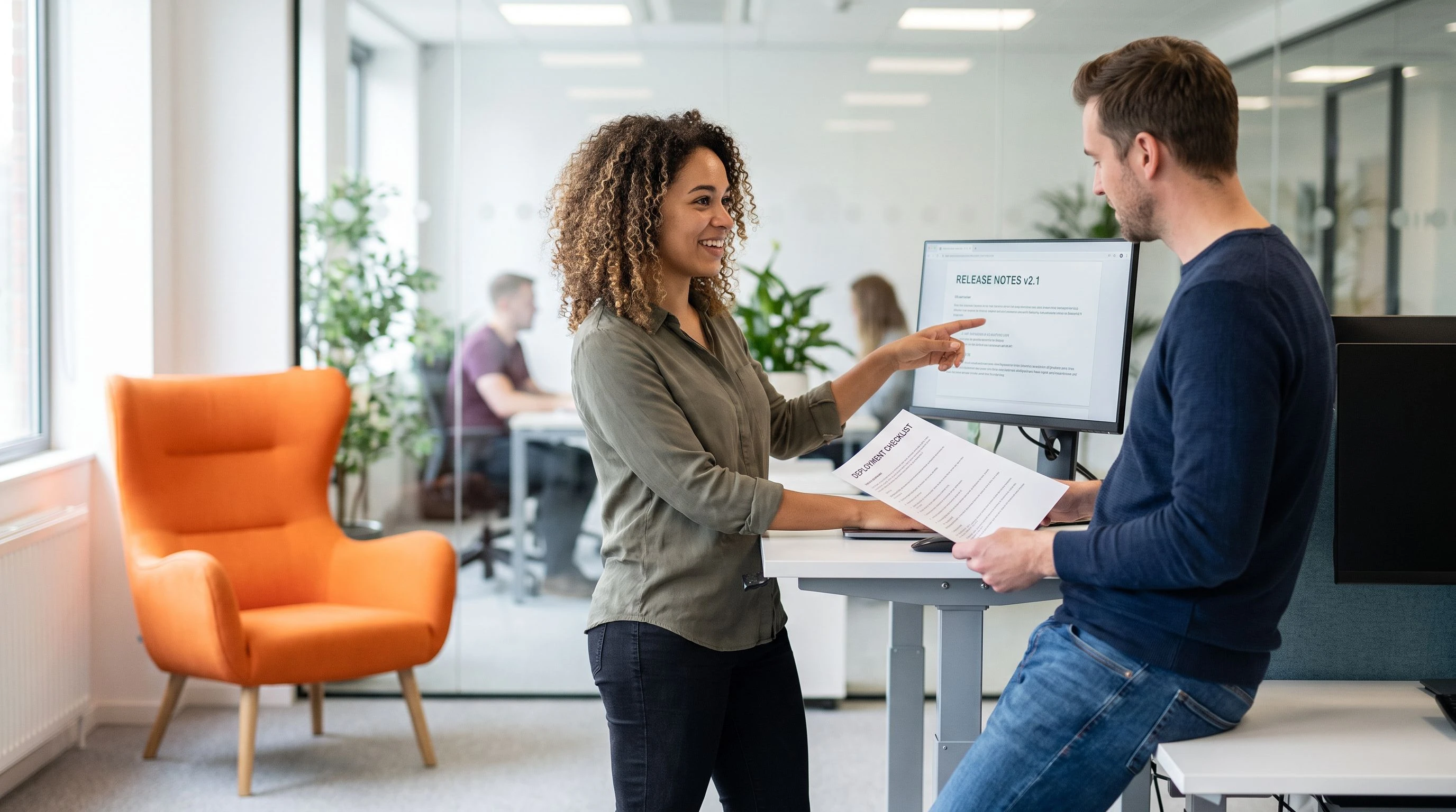 New iOS developer at a standing desk reviewing release documentation with a team lead, orange accent chair in the background