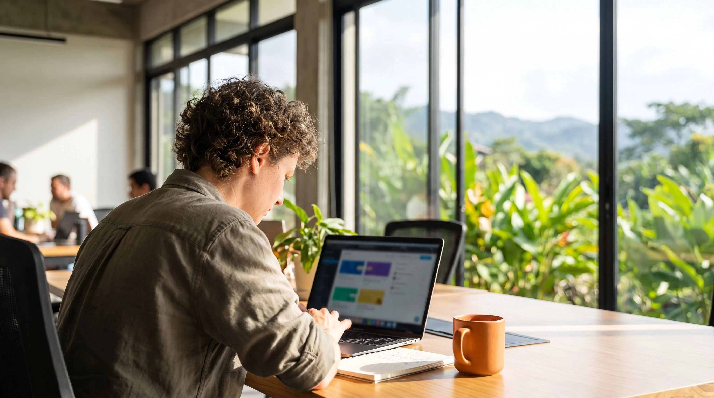 Nearshore LLM engineer reviewing model fine-tuning results on a laptop in a bright Costa Rica office, orange accent coffee mug on desk