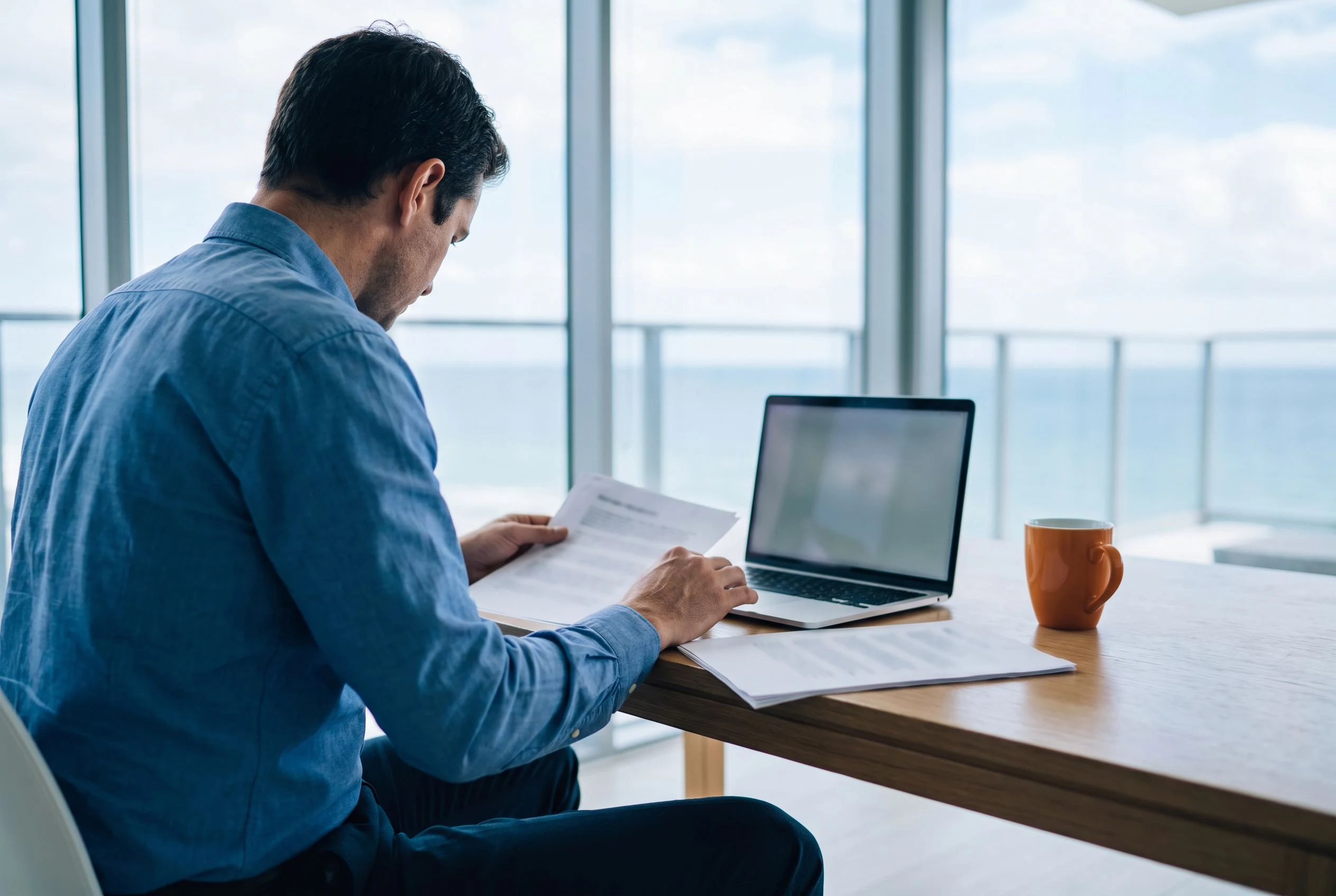Offshore accountant reviewing printed financial reports next to a laptop, orange coffee mug on desk, bright modern office