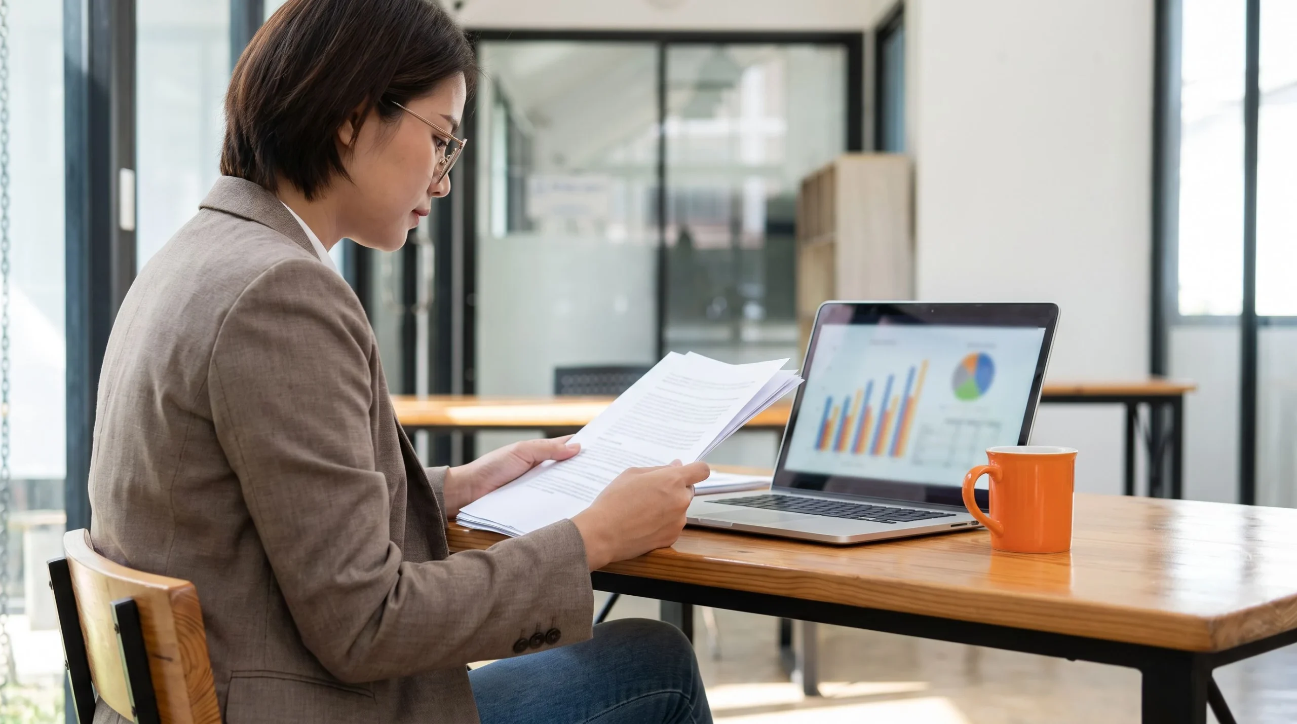 Finance professional reviewing offshore payroll salary benchmark documents beside a laptop with charts, orange coffee mug on desk