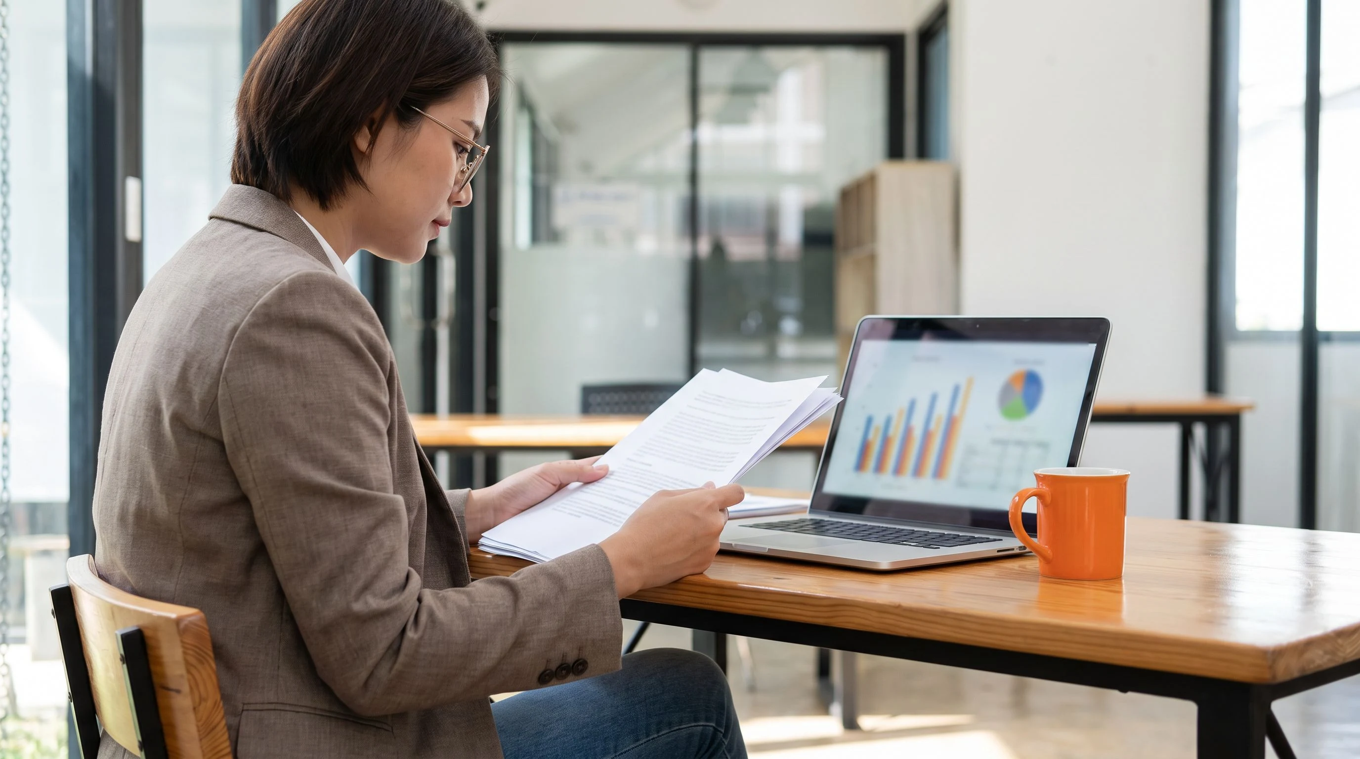 Finance professional reviewing offshore payroll salary benchmark documents beside a laptop with charts and an orange coffee mug