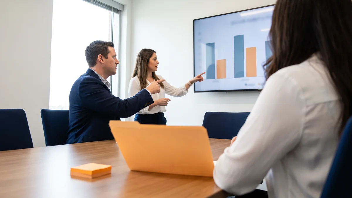 Two colleagues reviewing a help desk cost comparison on a wall-mounted monitor in a modern office